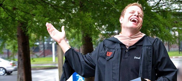 Business Administration Alumnus in graduation robe holding cap, standing outside under a tree laughing