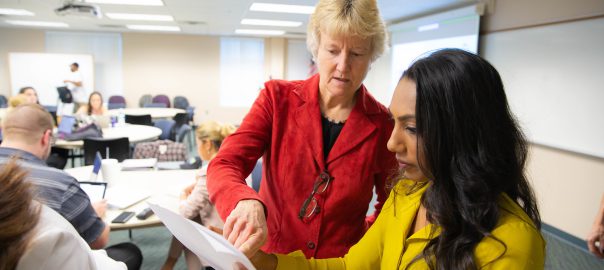 an instructor wearing a red blazer assists a student in a yellow dress, holding a paper