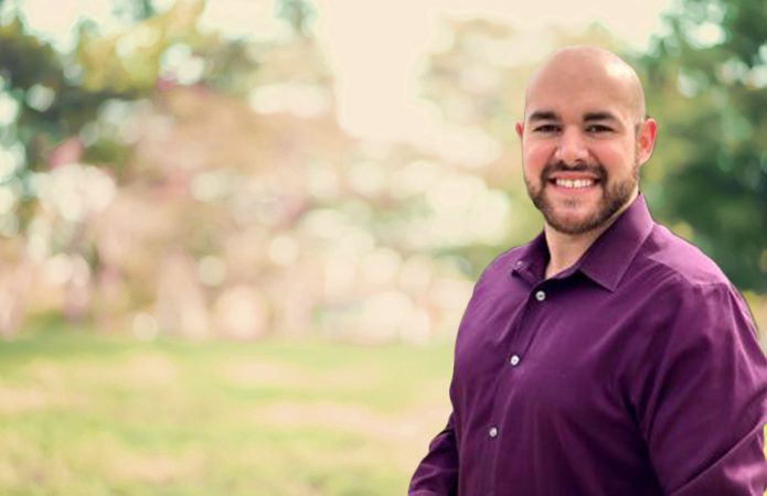 Gould, bearded and wearing a maroon button down shirt, open at the collar, smiles into the camera.