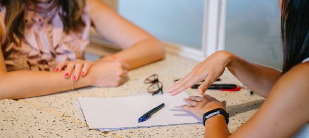 Two women going over paperwork for mock interview in an office setting. Two pens and a set of eyeglasses are placed on top of the paperwork.
