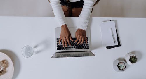 Individual working on a laptop at a desk with a notepad and coffee.