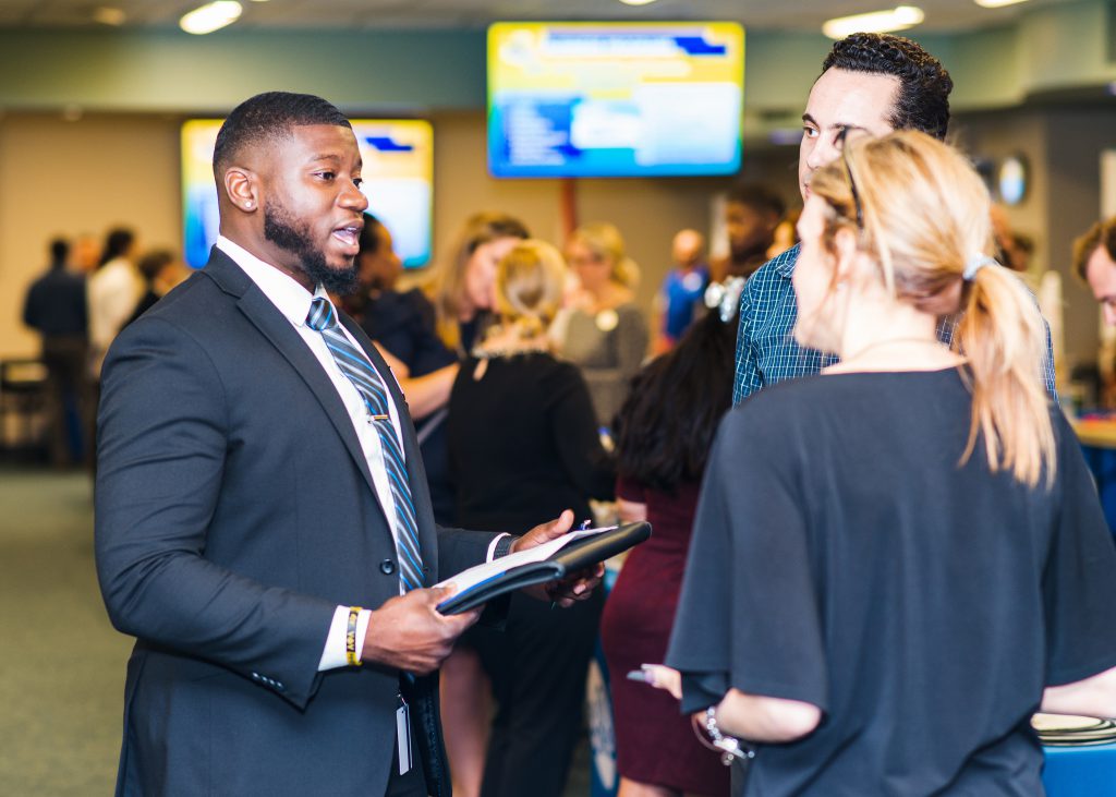Young man in a dark blue suit talks to prospective employers at a career fair.