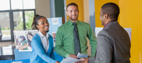Two students in business attire talk to a prospective employer at a career fair.