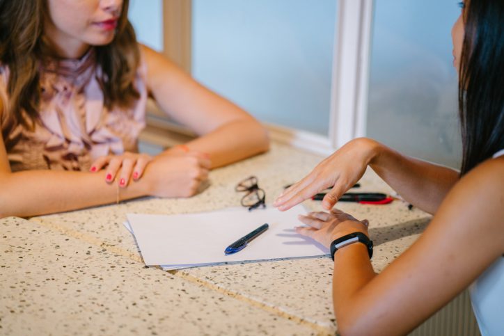 Two women going over paperwork for mock interview in an office setting. Two pens and a set of eyeglasses are placed on top of the paperwork.
