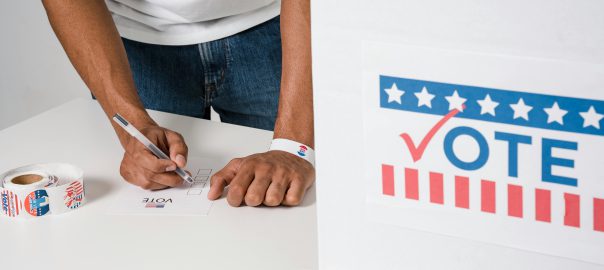 Person filling out an election ballot next to a ballot box that says "vote" on the front in red and blue lettering.