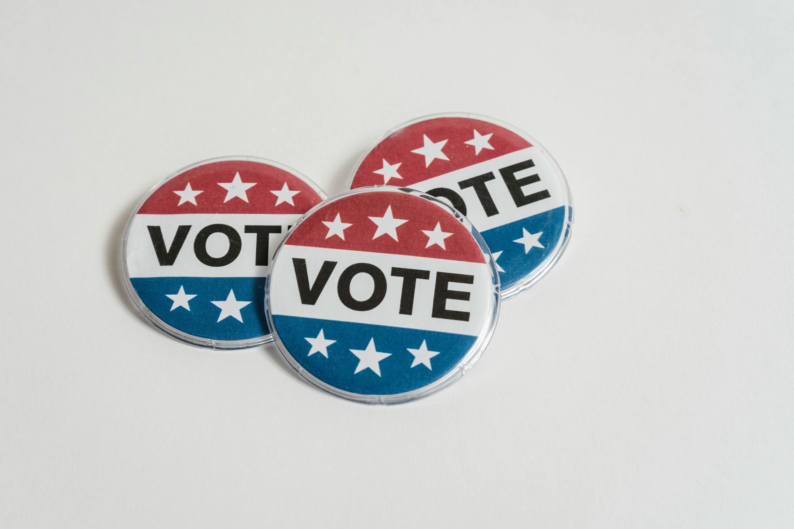 Three red, white and blue pins with the word Vote on them and six white stars are positioned on top of each against a white background.