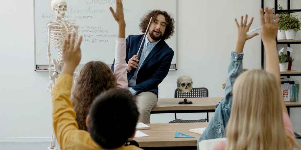 male teacher calls on a student in a room full of students with hands raised