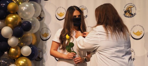 a nursing graduate wearing white scrubs and a black mask receives her nursing pin from her instructor