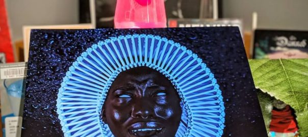 Neon Cover of a vinyl record in front of a red lava lamp.;