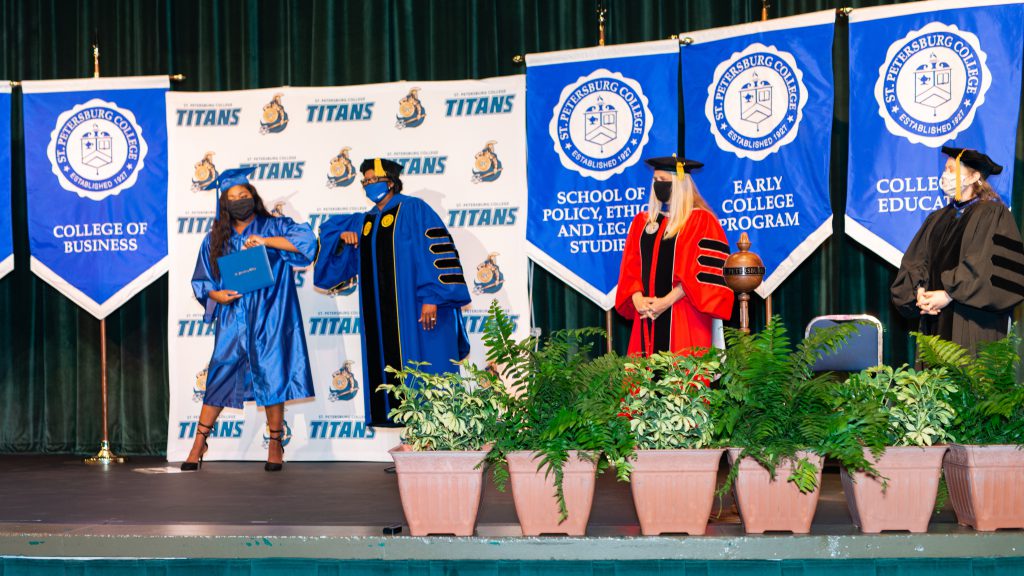 President Williams bumps elbows with a recent graduate on stage. Both are wearing face masks and blue graduation regalia.