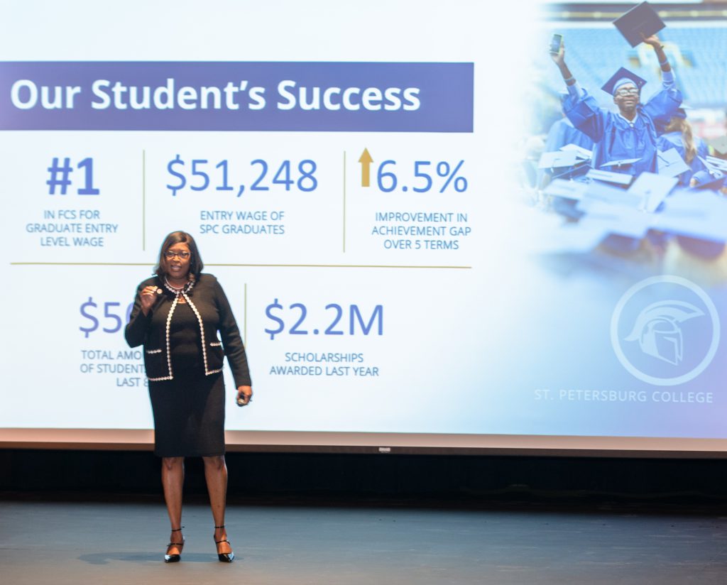 Dr. Williams speaks on stage in front of a screen displaying a powerpoint slide regarding student success.