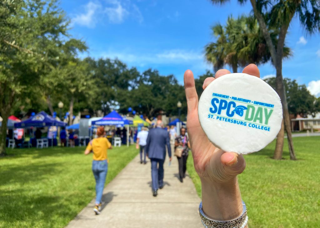 Underneath a bright blue sky, a person's hand holds up a cookie, covered in white frosting with the words SPC Day St. Petersburg College stamped in blue and green on top, in front of a group of tents, booths and people gathered for SPC Day 2022 at the St. Petersburg/Gibbs Campus.