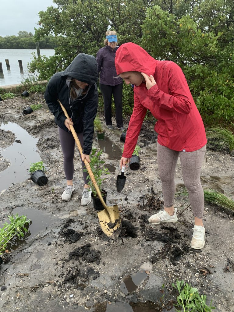 Two women, one in a black hoodie and one in a red hoodie, dig a hole in order to plant.