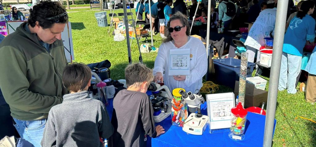 a woman stands behind a science exhibit table at an outdoor science festival, while a dad and his kids are seen from behind, watching her