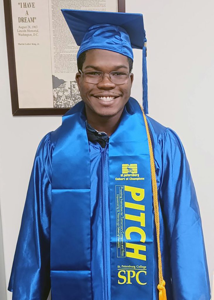 Young man in blue graduation gown and cap. The sash around his shoulders reads Pitch.